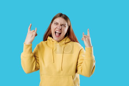 Young brunette 30s woman makes rock n roll sign isolated on blue background, studio portrait