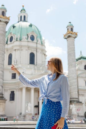 Young female tourist in sunglasses pointing finger near Catholic Church Karlskirche, Vienna. Saint Karl Cathedral. Traveling Europe in summer time. High quality photo