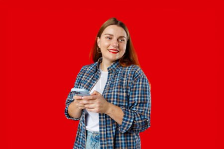 Lächelnde junge brünette Frau mit Smartphone, die isoliert auf rotem Hintergrund posiert, Studioporträt. Nette schöne junge Frau chattet per Telefon