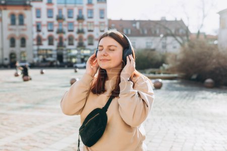 Happy young caucasian woman in good mood listening to music with headphones and standing on the street in the city. Music lover enjoying music.