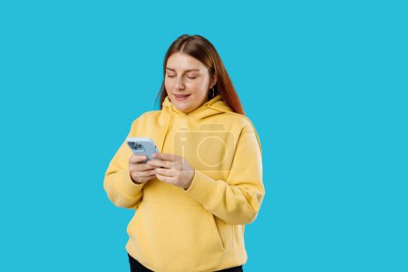 Lächelnde junge brünette Frau mit Smartphone posiert isoliert auf blauem Hintergrund, Studioporträt.