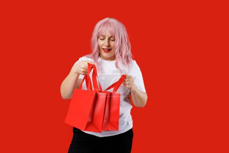 Porträt einer aufgeregten schönen Frau mit roten Einkaufstüten auf rotem Hintergrund. Black Friday, Sale, Buy Day Konzept. Pinkfarbene Haare, Studiodreh