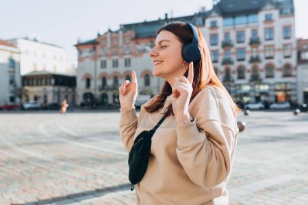 Happy young Caucasian woman in good mood listening to music with headphones and standing on the street in the city. Music lover enjoying music.