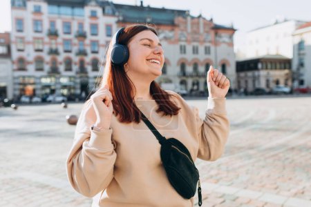 Happy young Caucasian woman in good mood listening to music with headphones and standing on the street in the city. Music lover enjoying music.