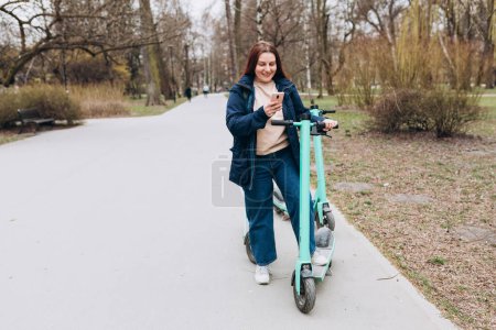 Lächelnde junge Frau, die neben einem Elektroroller mit Smartphone steht. Ökologischer Transport. Aktiver Lebensstil.