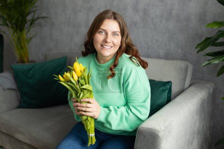Woman holding bouquet and siting on the sofa couch at home, hotel. 30s female rest and spend free spare time in living room indoors. People lounge or holiday concept