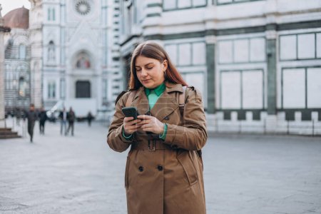 Photo for Woman walking on city street checks phone. 30s girl using smartphone near famous Duomo cathedral in Florence. Stylish women visiting Italian landmarks. Concept of travel, tourism and vacation in city - Royalty Free Image