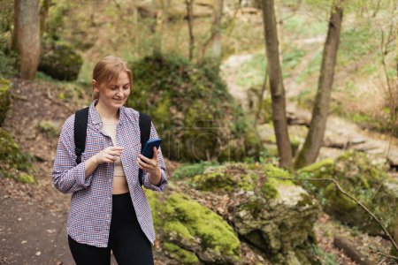 Téléchargez les photos : Fille touristique marche dans la forêt et en utilisant un smartphone. Jeune femme avec sac à dos randonnée sur la route dans la nature. Belle femme des années 30 marchant sur la nature en vêtements de sport - en image libre de droit