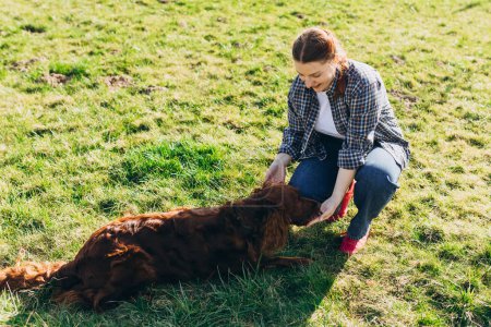 Cheerful happy young woman playing with her dog in the yard in summer. Beautiful Irish Setter dog is lying in grass