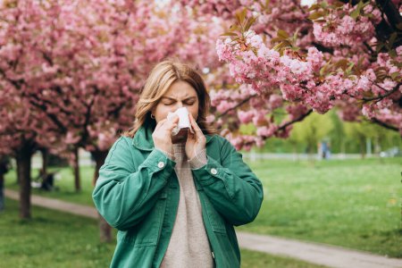 Jeune rousse éternuante avec essuie-nez parmi les arbres en fleurs dans le parc. Portrait de femmes malades éternue dans les tissus blancs, souffre de rhinite et le nez en cours d'exécution. Symptômes de rhume ou d'allergie.