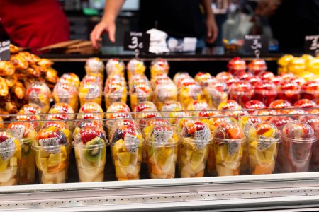 Téléchargez les photos : Salade de fruits disposée dans des tasses en plastique sur un marché. Fruits coupés et prêts à être vendus sur le marché. Commodité, mode de vie sain. Fraises, kiwi, melon, ananas, figues, dragon dans un verre plastique - en image libre de droit