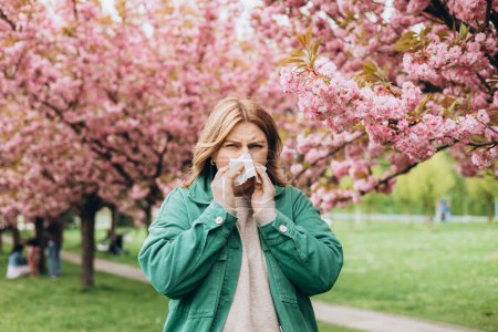 Jeune rousse éternuante avec essuie-nez parmi les arbres en fleurs dans le parc. Portrait de femmes malades éternue dans les tissus blancs, souffre de rhinite et le nez en cours d'exécution. Symptômes de rhume ou d'allergie.