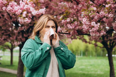 Sneezing young redhead woman with nose wiper among blooming trees in park. Portrait of sick women sneezes in white tissue, suffers from rhinitis and running nose. Symptoms of cold or allergy.