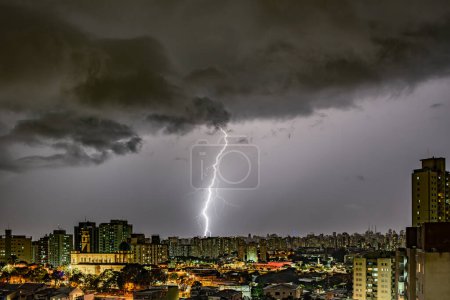 A dramatic color photo of a lightning storm over a cityscape at night. Bolts illuminate dark clouds and buildings, creating an intense atmosphere. City lights contrast with the ominous sky, showcasing nature's power.