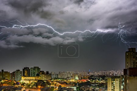 A dramatic color photo of a lightning storm over a cityscape at night. Bolts illuminate dark clouds and buildings, creating an intense atmosphere. City lights contrast with the ominous sky, showcasing nature's power.