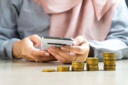femme d'affaires musulmane utilisant un smartphone ou une tablette avec un tas de pièces de monnaie à l'avant, symbole de la charia dividende d'investissement bancaire financier du concept de croissance des entreprises.