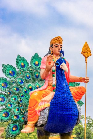 Estatua azul del Señor Siva sentado con una piel de tigre, sosteniendo un tridente y mostrando un gesto de bendición. Adornada con serpientes y cuentas, esta vibrante estatua se levanta majestuosamente contra un cielo despejado, encarnando el poder divino.