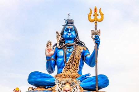 Foto de Estatua azul del Señor Siva sentado con una piel de tigre, sosteniendo un tridente y mostrando un gesto de bendición. Adornada con serpientes y cuentas, esta vibrante estatua se levanta majestuosamente contra un cielo despejado, encarnando el poder divino. - Imagen libre de derechos