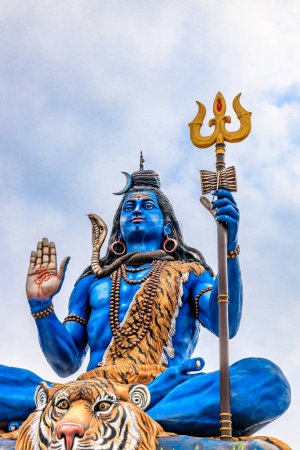 Estatua azul del Señor Siva sentado con una piel de tigre, sosteniendo un tridente y mostrando un gesto de bendición. Adornada con serpientes y cuentas, esta vibrante estatua se levanta majestuosamente contra un cielo despejado, encarnando el poder divino.