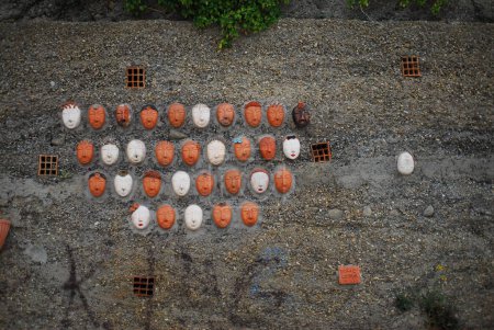 Photo for A view of an outdoor art installation featuring white and red masks arranged on a gravel surface. A group of clay heads, concrete art, a surrealist sculpture, Italy, Bussana Vecchia - Royalty Free Image