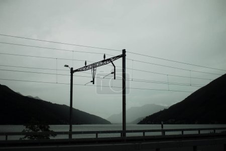 A serene railway platform with overhead lines is located near a calm lake, with misty mountains in the background