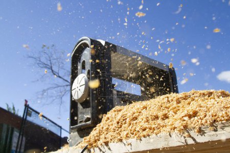 Sawdust is dispersed into the air as a 2x4 is making its way through a industrial sander on a construction site in daytime.
