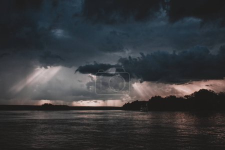 Ein atemberaubendes Bild, das das dramatische Zusammenspiel von Licht und Gewitterwolken über dem Sambesi-Fluss bei Sonnenuntergang einfängt, während Sonnenstrahlen durch den dunklen Himmel dringen und eine atemberaubende Szene schaffen.