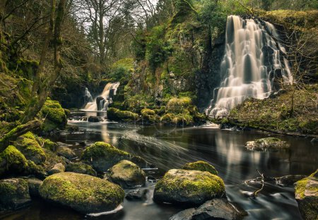 Langzeitbelichtungsaufnahme der Linn Jaw Wasserfälle in der Nähe von Livingston, Schottland, mit bemoosten Felsen im Vordergrund und um die Wasserfälle herum und weißen Schaumstreifen im Wasser. West Lothian. Großbritannien