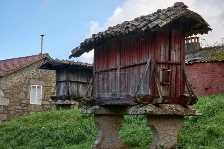 Vintage Horreo Kornspeicher auf Steinsäulen mit verwitterten roten Holzwänden unter einem bemoosten Ziegeldach in einer pastoralen Umgebung