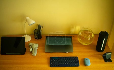 Un espacio de trabajo de alta tecnología, completo con un ordenador portátil, tableta, teclado, ratón, lámpara y una pecera, que refleja la vida de un estudiante de informática dedicado.