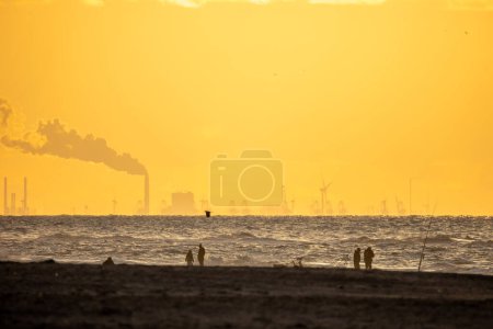 People walk on the shores of the North Sea. Beautiful orange, yellow sunset. The sea is in the background. Beautiful storm and waves. Yellow sand in the foreground.