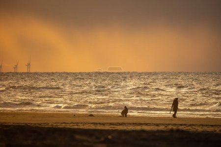 Les gens marchent sur les rives de la mer du Nord. Belle orange, coucher de soleil jaune. La mer est en arrière-plan. Belle tempête et vagues. Sable jaune au premier plan.
