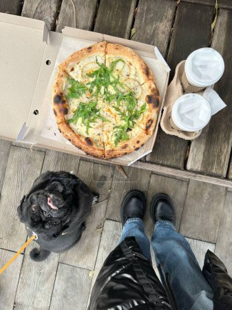 pizza, two disposable glasses of coffee on the wooden table,  human  legs, small black dog