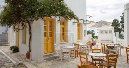 Grecia. Isla de Tinos Cícladas. Café tradicional al aire libre con ventanas amarillas en el pueblo de Pyrgos. Silla y mesa vacías en el patio pavimentado