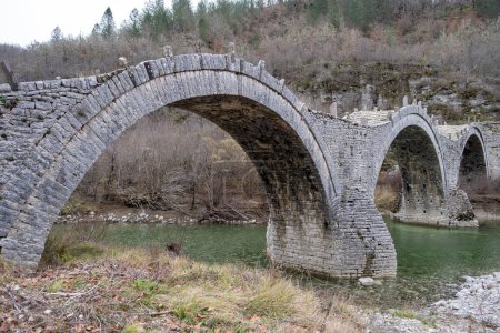 Photo for Kalogeriko or Plakidas ancient stone bridge with three arch over Voidomatis river water, Zagoria Epirus Greece. Dry tree background. - Royalty Free Image