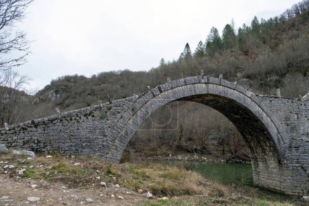 Photo for Kalogeriko or Plakidas ancient stone bridge. View of one of three arch over Voidomatis river water, Zagoria Epirus Greece. Dry tree background. - Royalty Free Image
