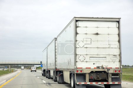 Photo for Three trailer road train transporting commercial cargo in USA highway. White long heavy vehicle rear, travels with cloudy sky background. - Royalty Free Image