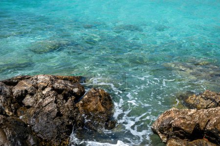 Vor klarem, transparentem, türkisfarbenem Meerwasserhintergrund bricht schäumende Welle an Felsen. Griechenland Agistri Insel Aponisos Strand. Ansicht von oben