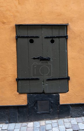 Ventana de madera cerrada gris oscuro en la pared de color naranja, Stockholm Sweden Old Town. Ventana vintage con agujero y pestillo se ve como una mala cara. Vertical
