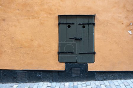 Dark grey closed wooden window on orange color wall, Stockholm Sweden Gamla Stan Old Town. Vintage window with hole and latch looks like a bad face.