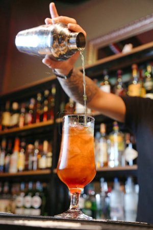 A bartender expertly pours a colorful cocktail into a stemmed glass, holding a shaker in one hand.