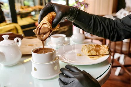 A person wearing black gloves carefully pours a cup of coffee from a ceramic coffee pot.