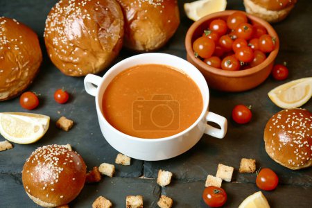 A cup of tomato soup surrounded by bread and cherry tomatoes, presenting a comforting and appetizing meal.