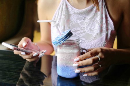Téléchargez les photos : Une femme est assise à une table, tenant un pot de liquide tout en utilisant un téléphone portable. - en image libre de droit