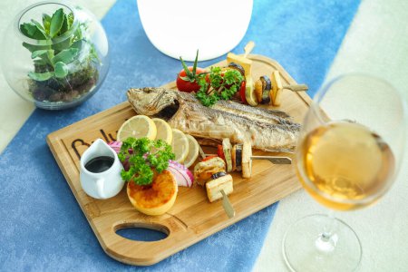 Téléchargez les photos : Une planche à découper en bois garnie d'un poisson fraîchement préparé servi avec un verre de vin. - en image libre de droit