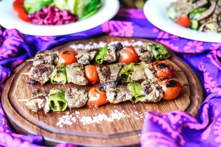 Eine Holztafel mit verschiedenen Fleisch- und Gemüsesorten, darunter gegrilltes Steak, Huhn, Wurst, Paprika, Zwiebeln und Zucchini.