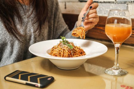 Téléchargez les photos : On voit une femme déguster une assiette de spaghettis en la mangeant à la fourchette, accompagnée d'un verre de jus d'orange rafraîchissant.. - en image libre de droit