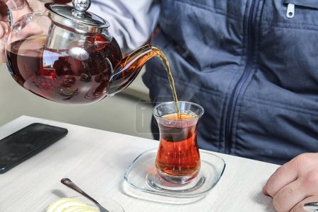 A person pours tea from a teapot into a glass cup on a wooden table.