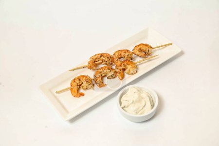 Une assiette blanche garnie de succulentes brochettes de crevettes placées à côté d'un bol rempli d'une trempette savoureuse.