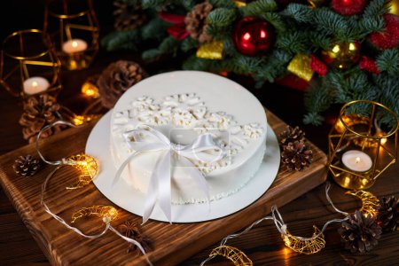 Foto de Pastel de mousse blanco navideño decorado con copos de nieve y un lazo en el fondo de velas, guirnaldas, ramas de abeto y conos - Imagen libre de derechos